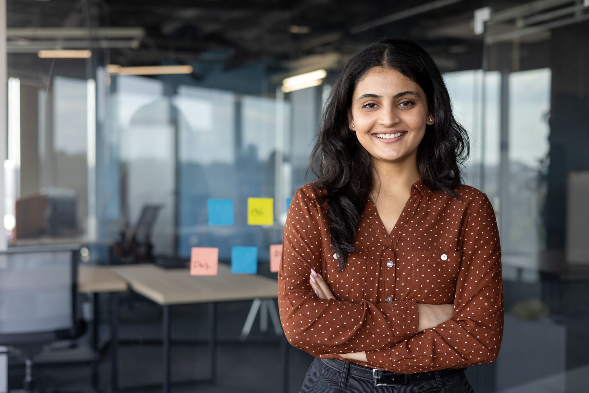 Successful confident businesswoman at workplace inside office, woman with crossed arms smiling and looking at camera, standing near window. Satisfied office worker of company